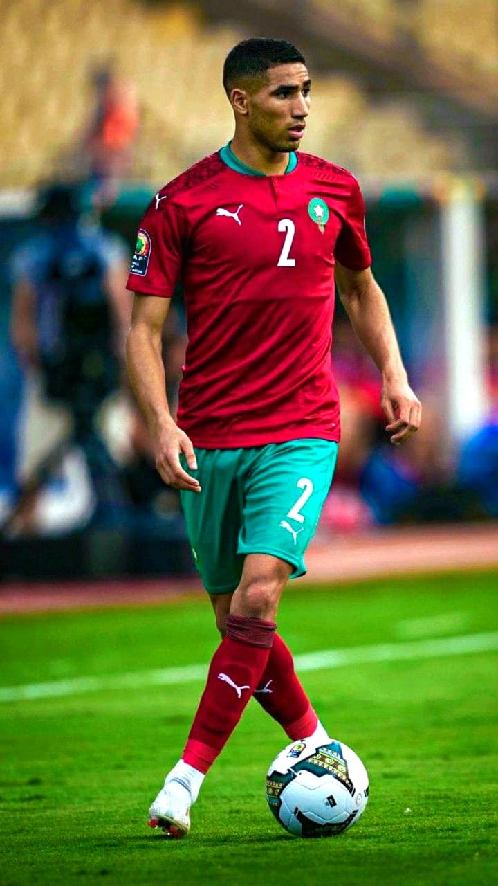 A soccer player in a red and green Morocco jersey with number 2 controls the ball on a grassy field during a match, as fans tune in from around the world via Colombia IPTV.